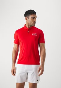 Red polo shirt made of lightweight fabric, featuring a three-button placket and embroidered logo on the chest. Paired with white shorts.