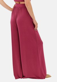 Široké kalhoty v barvě červené krve s elastickým pasem, hladkou texturou a splývavým designem. Na nohou jsou vidět sandály na pásky.