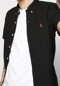 Svart knappad skjorta med korta ärmar, rund krage, ljusa knappar och en liten röd logga på bröstet. Bärs över en vit t-shirt.