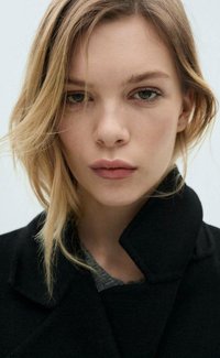 Črn plašč z visokim ovratnikom, mehkega otipa, nošen preko temno sive srajce. Model ima slojevite blond lase in zaprta usta. Nevtralen ozadje.