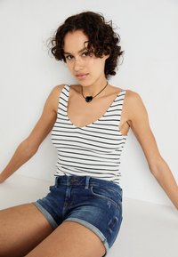 White ribbed tank top with navy stripes, paired with blue denim shorts featuring rolled cuffs and slight distressing. Black heart necklace accent.