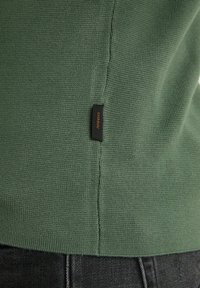 Pull en maille vert avec une petite étiquette noire "CHASIN" près de la couture latérale, associé à un jean en denim foncé.