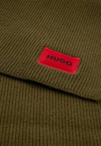 Tessuto a maglia verde a costine con un'etichetta rossa che riporta la scritta "HUGO" in nero. La texture è morbida e il design è semplice e contemporaneo.