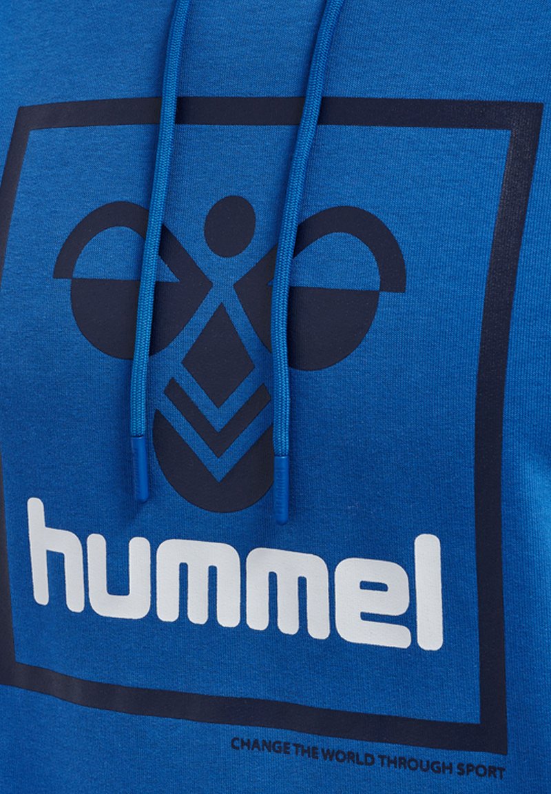 Blauwe stof met zwart geometrisch bijlogo en witte "hummel"-tekst binnen een zwart vierkant, met daaronder "VERANDER DE WERELD DOOR SPORT".