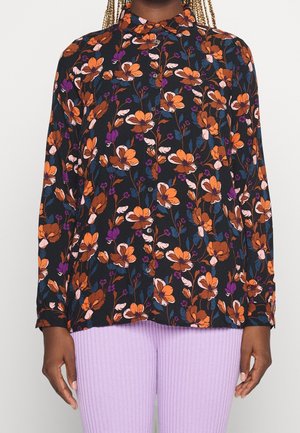 Personne portant une chemise à manches longues boutonnée à motif floral sombre avec des fleurs orange, violettes et roses, et un pantalon côtelé violet clair.