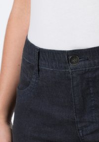 Donkerdenim broek met een elastische tailleband, voorkwaktes en een metalen knoopsluiting; gladde textuur met subtiele schaduwen.