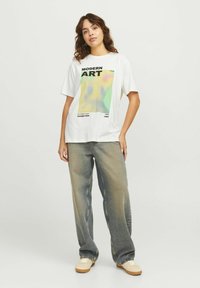 Camiseta de algodón blanca con un gráfico de "Arte Moderno" en colores suaves, combinada con unos pantalones vaqueros azules desgastados y suéteres beige.