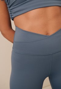 Legginsy z wysokim stanem w stonowanym niebieskim kolorze, wykonane z gładkiego, elastycznego materiału z płaskimi szwami i talią w kształcie litery V.