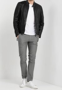 Giacca in pelle nera con spalle imbottite, chiusura con zip, indossata sopra una t-shirt bianca, abbinata a pantaloni grigi aderenti e scarpe da ginnastica bianche.