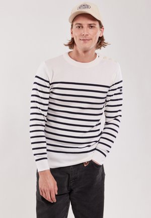 Pull blanc avec des rayures horizontales marines, col rond et accents de boutons sur l'épaule ; associé à un pantalon sombre et une casquette beige.