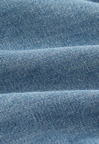 Denim-kangas vaaleansinisenä, jossa on teksturoitu, kudottu kuvio. Useat laskokset luovat pehmeän roikkuvuden, joka esittelee hieman vaihtelevaa värigradienttia.