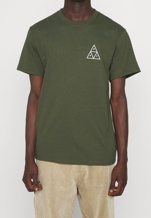 Mann in olivgrünem Kurzarm-T-Shirt mit weißem dreieckigen Logo und beigen Cordhosen vor einem schlichten weißen Hintergrund.