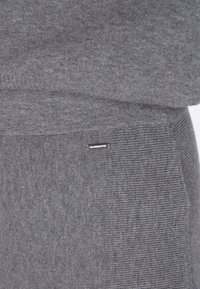 Tissu tricoté gris avec une texture côtelée et une petite étiquette en métal. Le design est simple, sans motifs ni embellissements distincts.