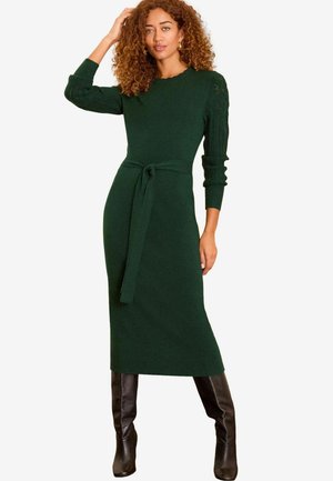 Rochie verde tricotată cu un corp ajustat și mâneci lungi, texturate. Dispune de un talie legată și se oprește la mijlocul gambei. Poartă cizme negre până la genunchi.