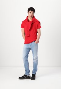 Rode katoenen T-shirt met korte mouwen, gecombineerd met lichtblauwe spijkerbroek. Een rode sweatshirt om de nek en zwarte sneakers.