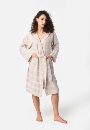 Woman with curly hair wearing a beige striped bathrobe tied at the waist, standing barefoot against a plain white background.