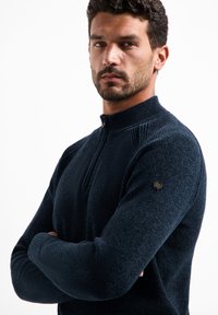 Navyblauwe zip-up trui met textuur breisel, hoge kraag en geribde manchetten. Heeft een klein zwart logo accent op de linkermouw.