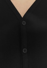 Schwarze Rippstrickjacke mit zwei schwarzen Knöpfen, die geschlossen ist und einen kleinen Teil der Haut am Hals zeigt.