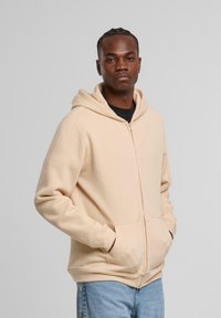 Beige zip-up hættetrøje med snoretræk, forlommer og ribbede manchetter, båret over en sort skjorte og parret med lysblå jeans.