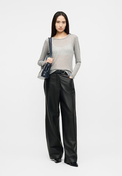 Žena oblečená v priehľadnom sivom top s dlhými rukávmi, čierne kožené nohavice s širokými nohavicami, čierny opasok, čierne lodičky a nesúca čiernu kabelku na ramene.