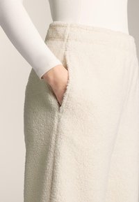 Puha fehér anyagból készült polárnadrág, melynek felülete texturált, oldalzsebekkel rendelkezik, és laza szabású, látható varrásokkal a szegélyeken.