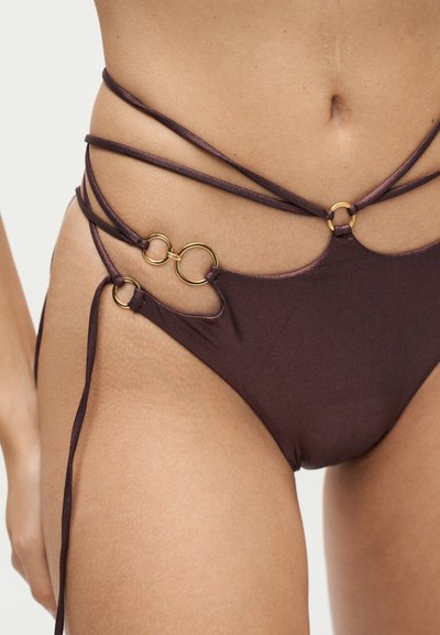 Gros plan d'une femme portant un bas de bikini marron avec des fines bretelles et des détails en anneaux dorés autour de la taille et de la hanche.