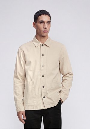 Mand iført en beige knap-op jakke med to forlommer og sorte bukser, stående mod en ensfarvet lys baggrund.