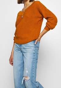 Gestrickter oranger Pullover mit weitem Ausschnitt und lockerem Schnitt, mit gerippten Bündchen und Saum, kombiniert mit hellblauen, abgenutzten Jeans.