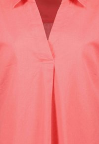 Blusa color corallo con scollatura a V, maniche corte e dettaglio plissettato frontale. Realizzata in tessuto leggero con una texture liscia.