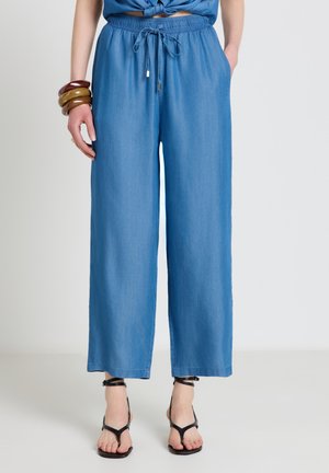 Femme portant un pantalon bleu ample à cordon avec poches, des sandales noires à brides, et des bracelets en bois au poignet gauche, debout devant un fond uni.