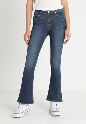 Kobieta ubrana w ciemnoniebieskie dzwony z wysokim stanem i białe trampki, stojąca na tle jednolitego jasnego tła.