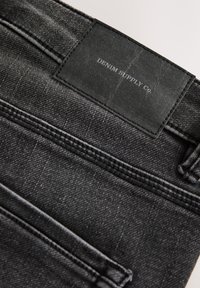 Primo piano della tasca posteriore di un paio di jeans in denim nero con etichetta in pelle nera recante la scritta "DENIM SUPPLY Co." e dettagli di cucitura visibili.