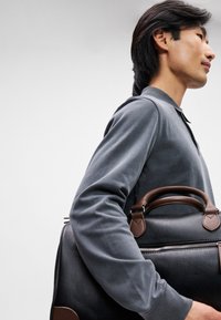 Μαύρη υφασμάτινη τσάντα με καφέ δερμάτινες λεπτομέρειες, με κλείσιμο με φερμουάρ και διπλές λαβές, κρατημένη από ένα άτομο με γκρι μακρυμάνικο πουκάμισο.