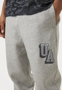 Grå sweatpants lavet af blødt stof har en struktureret overflade, sidelommer og et fremtrædende "UA" logo i mørkegrå på venstre lår.