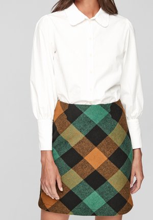 Žena v bílé košili s dlouhými rukávy na knoflíky s nadýchanými rukávy a v sukni s vysokým pasem v kostkovaném vzoru v zelených, oranžových, černých a hnědých odstínech.