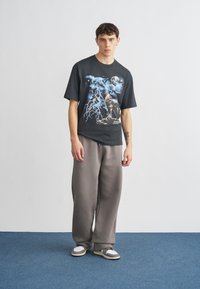 Schwarzes grafisches T-Shirt mit markanter blau-weißer Illustration, kombiniert mit locker sitzenden grauen Jogginghosen und lässigen Turnschuhen mit Akzenten.