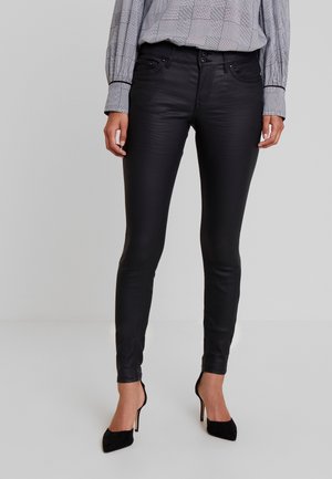 Woman wearing black leather skinny pants, black pointed high heels, and a black-and-white checkered long-sleeve shirt against a gray background.