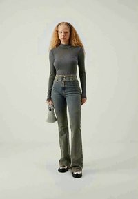 Mlada ženska z kodrasto pričesko nosi sivo rebrasto turtleneck, svetlo jeans kroje visokega pasu, črne čevlje z metalnimi konicami in drži majhno sivo torbico.