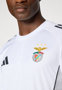 Fehér sportpóló fekete Adidas csíkokkal, színes Benfica logóval, fölötte egy sas és három csillag található.