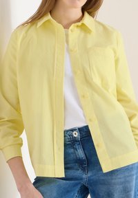 Femme portant une chemise en velours côtelé jaune partiellement boutonnée sur un haut blanc, associée à un jean denim bleu.