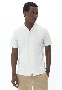 Korte mouwen, met kraag, lichtgrijze shirt van stof met een knoopsluiting aan de voorkant en een subtiele textuur; gecombineerd met beige broek.