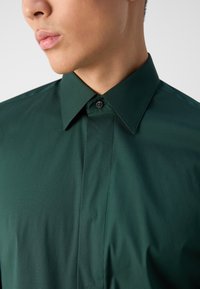Camisa verde oscura con cuello clásico, un ligero brillo y un botón negro en la tapeta. Tejido suave con un corte entallado.