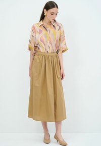 Blusa leggera e multicolore con motivi astratti; gonna plissettata beige fino al ginocchio; scarpe con tacco neutre; orecchini circolari.