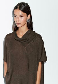 Brun stickad topp med draperad halsringning, korta ärmar och en lös passform. Texturerat tyg med subtila detaljer. Modellen poserar mot en enfärgad bakgrund.