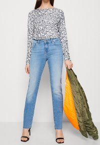 Top à manches longues imprimé léopard, jeans bleu clair slim, et sandales à talons noires. Modèle tenant une veste verte et orange.