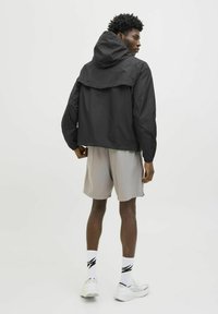 Zwarte lichte jas met capuchon, met lange mouwen en een ontspannen pasvorm, gecombineerd met grijze shorts en witte sportsokken.