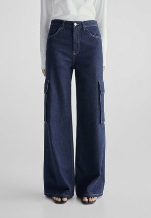 Jean cargo bleu foncé à jambes larges avec poches latérales, fermeture par bouton et fermeture éclair, porté avec un haut blanc à manches longues et des sandales noires ouvertes à l’avant.