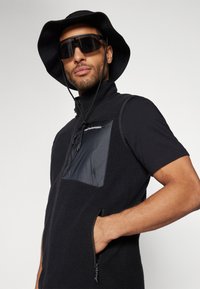Gilet en polaire noire avec une fermeture éclair à l'avant, poche sur la poitrine en tissu lisse et poches latérales à fermeture éclair. Porté avec un chapeau noir à large bord et des lunettes de soleil.