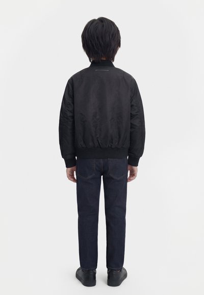Παιδί που στέκεται γυρισμένο πλάτη, φορώντας μια μαύρη μπουφάν τύπου μπόμπερ, σκούρο μπλε τζιν και μαύρα αθλητικά παπούτσια σε απλό λευκό φόντο.