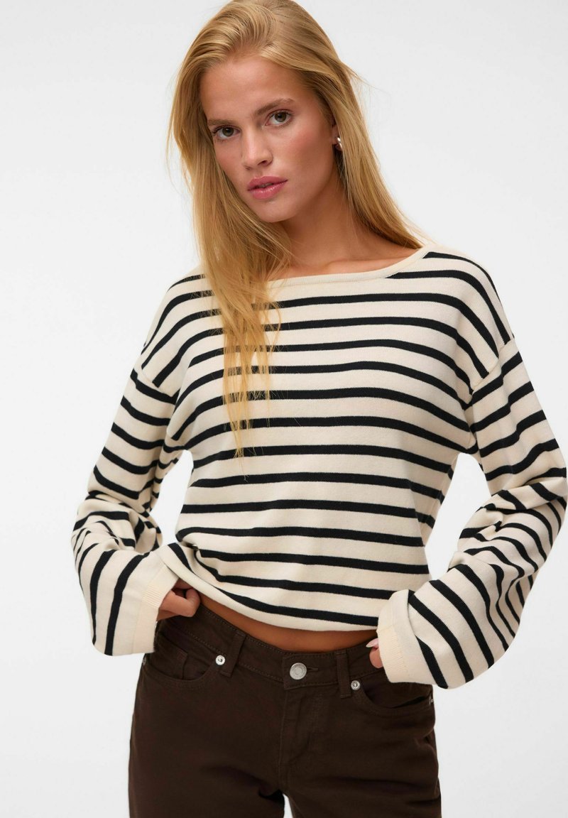 Femme blonde portant un haut à manches longues crème et noir rayé et un pantalon marron foncé, les mains dans les poches avant.